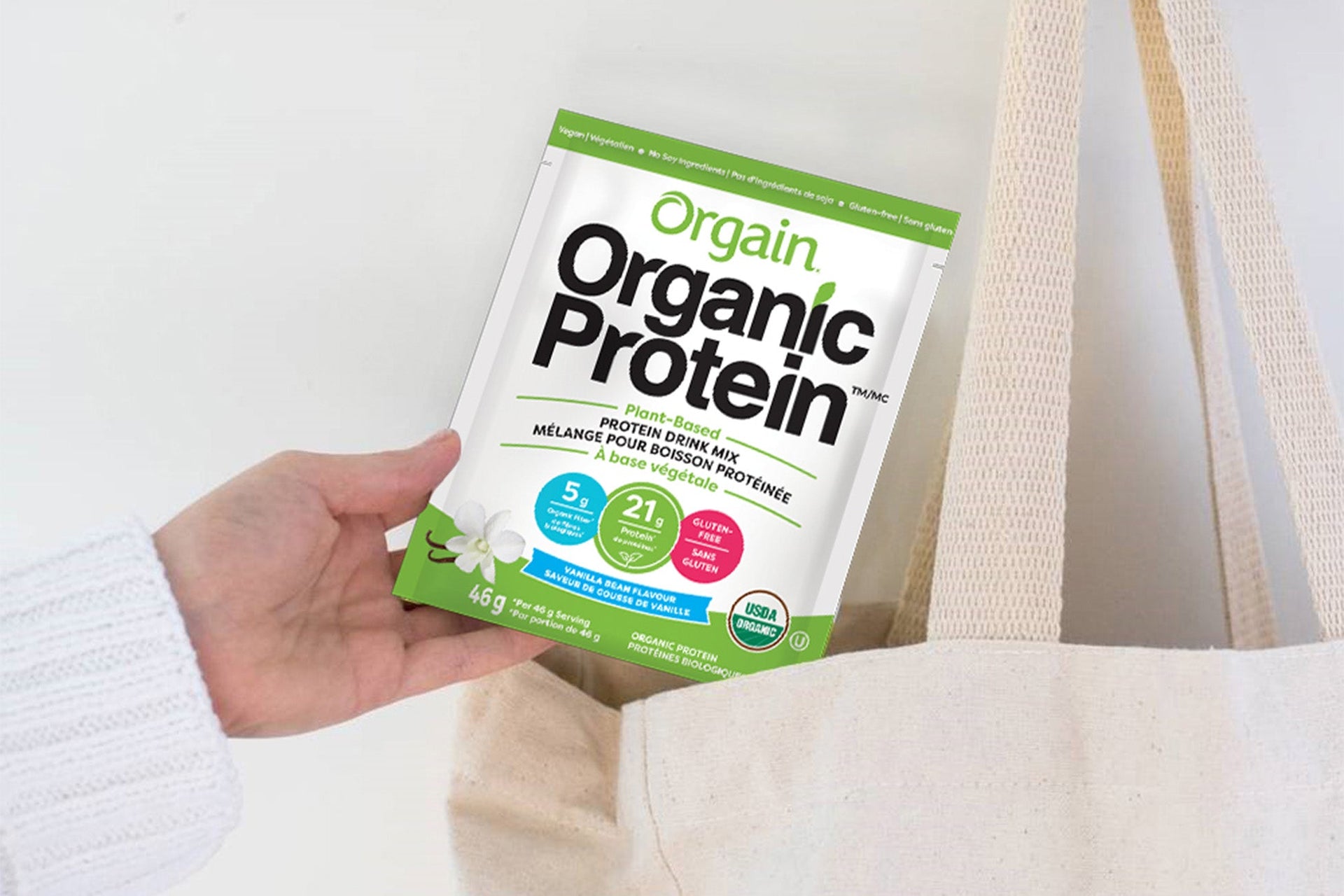 A hand holds a packet of Orgain Organic Protein Powder near an open canvas bag, highlighting 21g protein, gluten-free, and vegan.