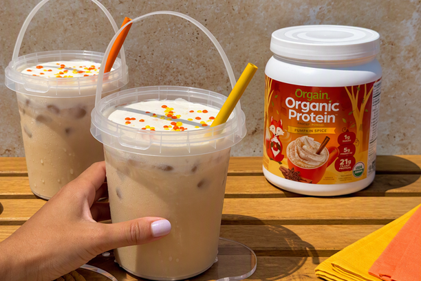 Two iced pumpkin spice protein lattes served in clear bucket-style cups with handles, topped with whipped cream and fall-colored sprinkles, next to a container of Orgain Pumpkin Spice Organic Protein powder.