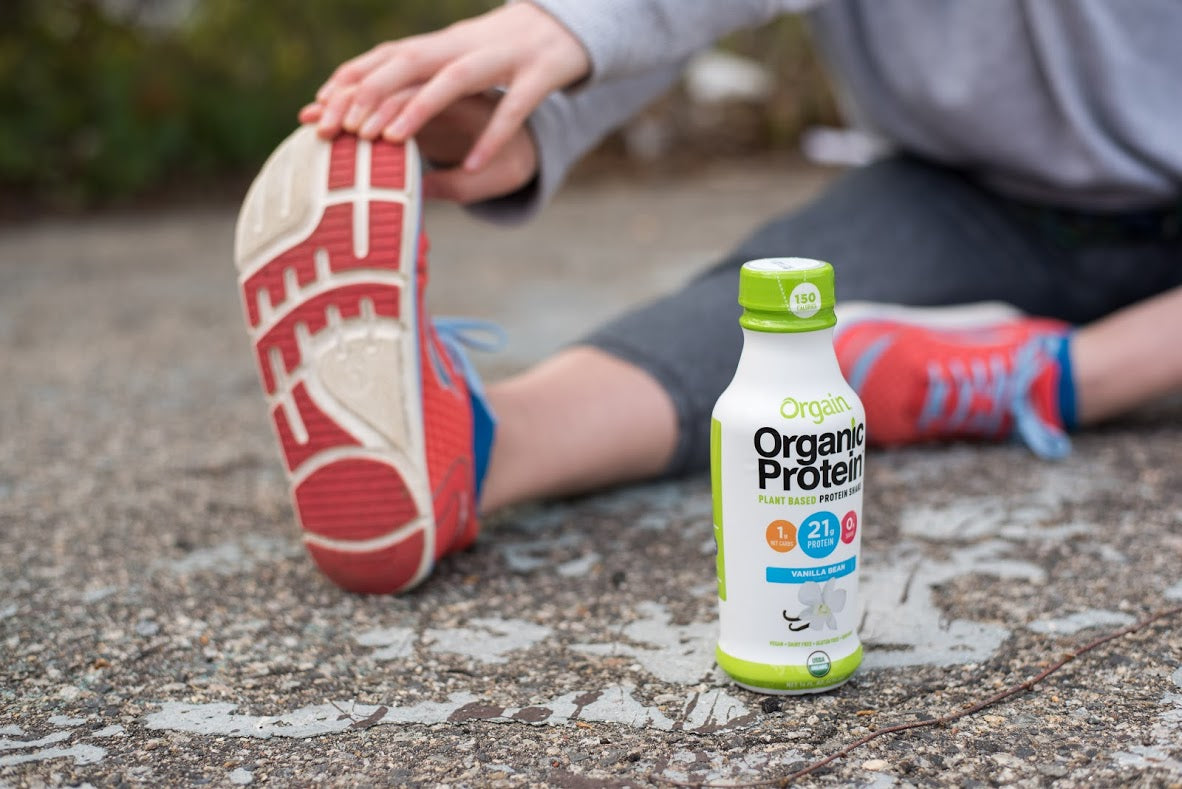 A person in athletic wear stretches one leg with their foot extended forward. A bottle of Orgain Organic Protein shake with vanilla flavor stands on the ground beside them. The background is slightly blurred, showing an outdoor setting.