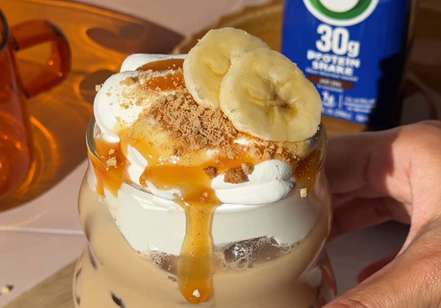 Close up of iced banana cream pie protein latte topped with whipped cream, caramel drizzle, crushed cookie crumbs, and banana slices, with an Orgain 30g Protein Shake in the background.