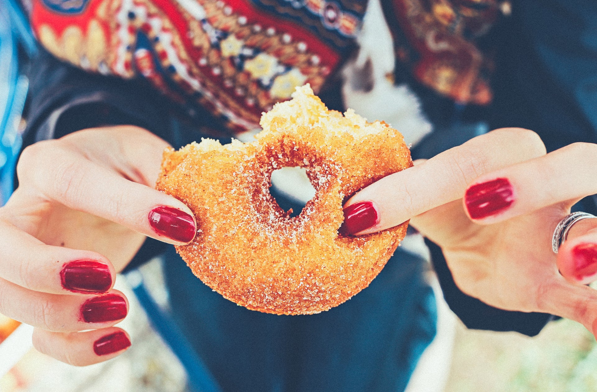 A person with red-painted nails holds a bitten sugar-coated donut. The person is wearing colorful clothing, and only their hands are visible in the image. The background is slightly out of focus, emphasizing the donut and the hands.