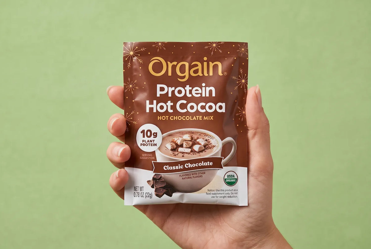 A hand holding a single packet of Orgain Protein Hot Cocoa Classic Chocolate Mix against a solid light green background.