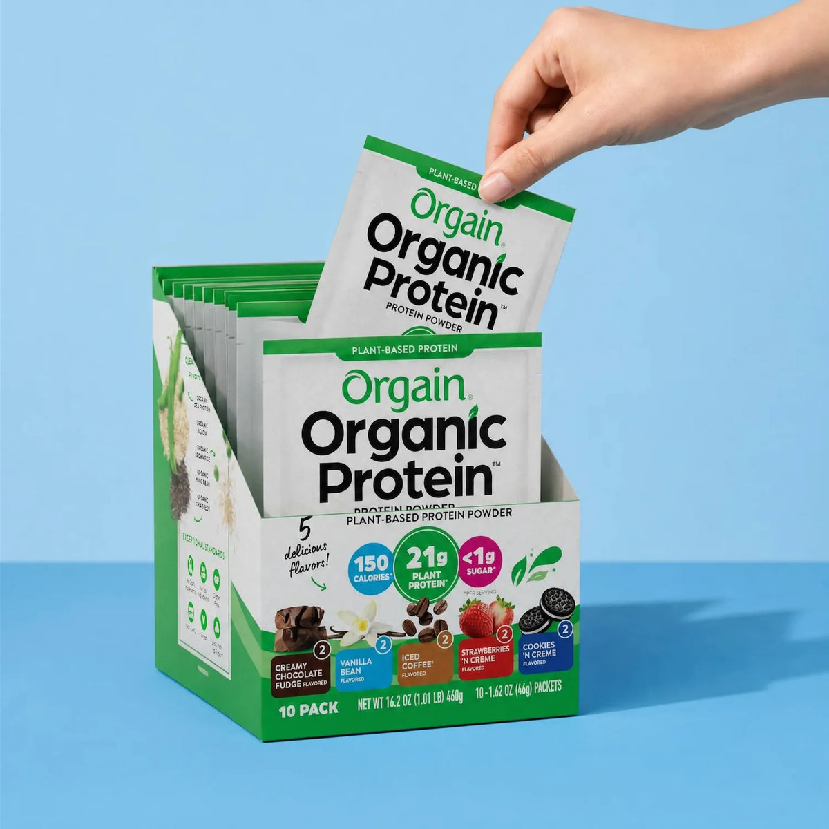 Hand removing a single-serve packet from an Orgain Organic Protein Plant-Based Protein Powder Variety Pack display box containing assorted flavors.