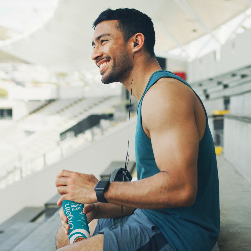 A smiling man in a teal tank top sits in a stadium holding a green and white Organ protein powder bottle, wearing earbuds and a smartwatch.
