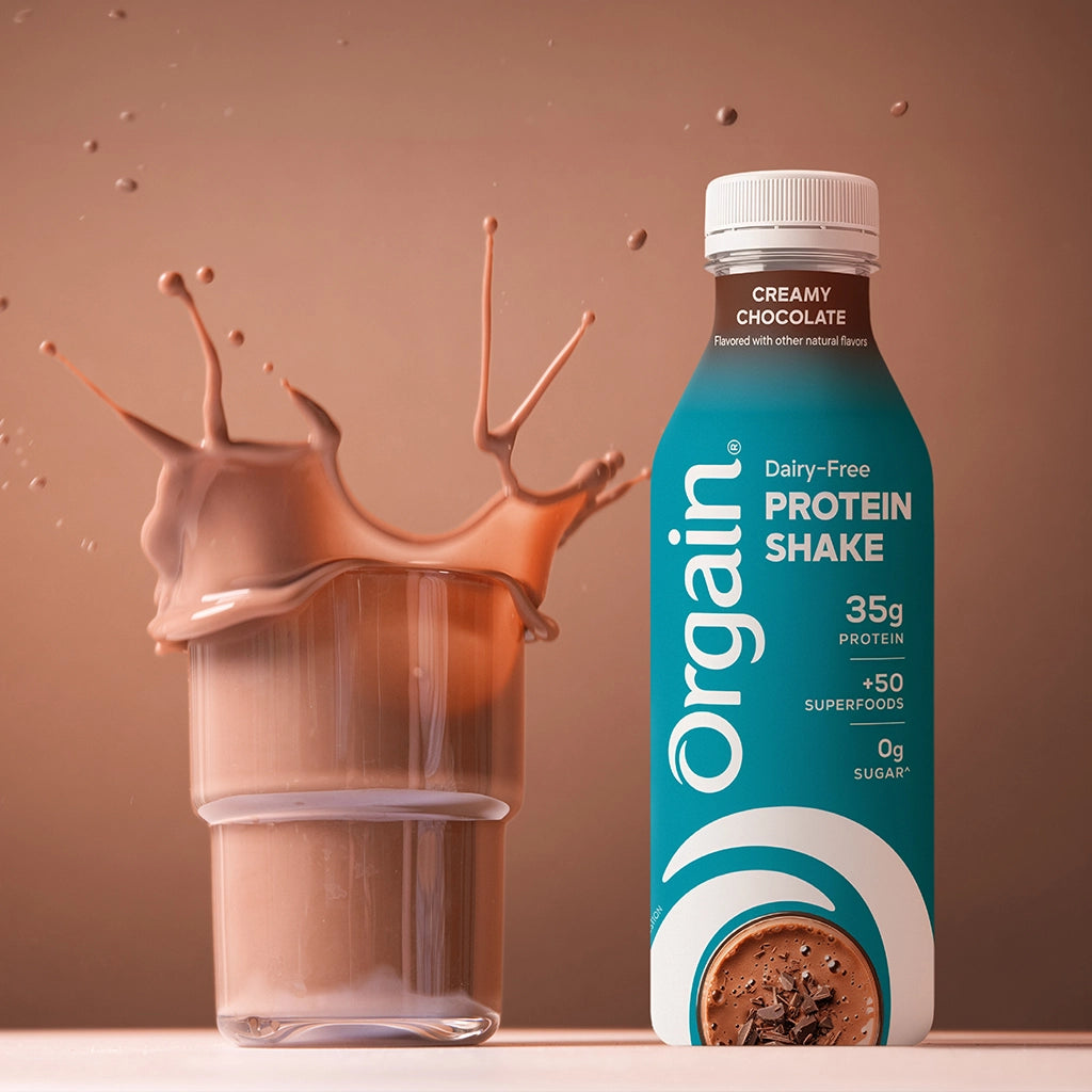 An Orgain protein shake bottle labeled Creamy Chocolate stands next to a glass of chocolate protein shake with a dramatic splash effect, set against a warm brown studio background.