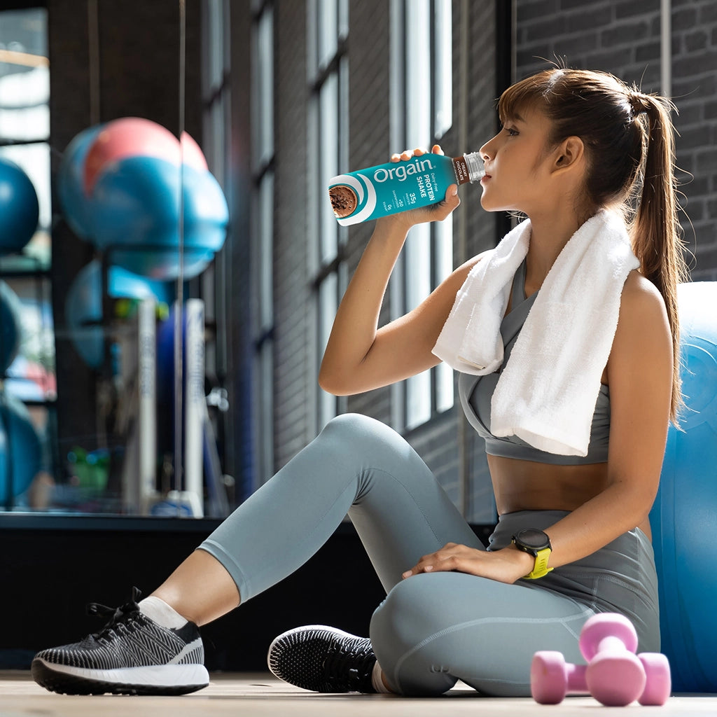 A woman in a gym setting sits on the floor, drinking from a blue Orgain protein powder bottle while wearing a gray sports bra and leggings with a white towel over her shoulder.